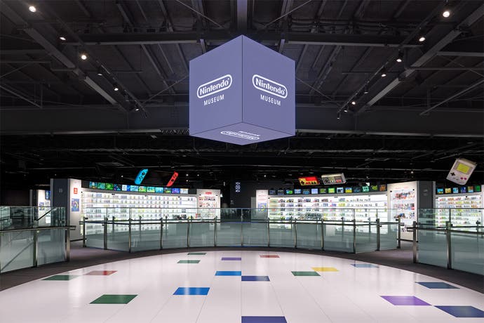 An interior photograph of the second floor of the Nintendo Museum's Product Floor exhibit, showing a white oval floor, a cube suspended in the ceiling with the Nintendo Museum logo on all sides, and two exhibit walls dedicated to the Switch and Famicom.