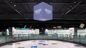 An interior photograph of the second floor of the Nintendo Museum's Product Floor exhibit, showing a white oval floor, a cube suspended in the ceiling with the Nintendo Museum logo on all sides, and two exhibit walls dedicated to the Switch and Famicom.