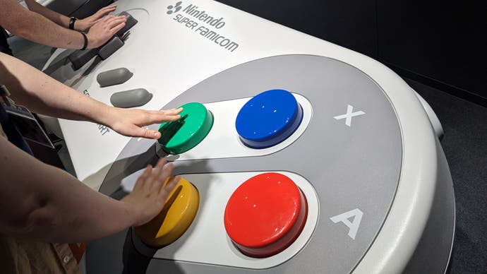 A close up photograph of two people pressing giant buttons on a giant SNES controller in the Nintendo Museum.