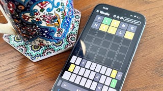 A photo of an iPhone and cup of tea on a table. An in-progress game of Wordle is visible on the phone's screen.