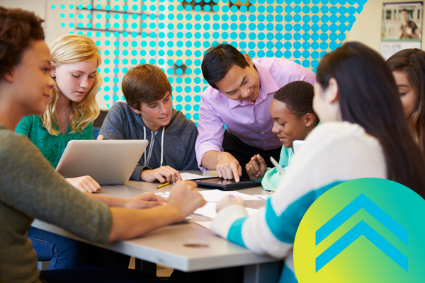 High school students are sitting in a classroom practicing math on devices. A teacher points to a particular part of the tablet screen while he instructs one of the students.